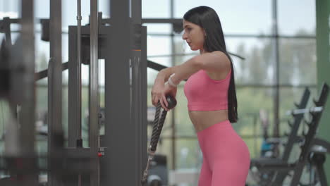 a hispanic brunette woman in a pink suit pulls a rope in a crossover with her hands to train her shoulders. shoulder workout in a trainer. professional woman instructor