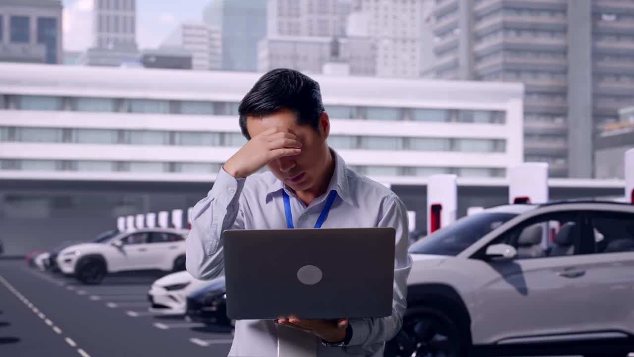 近代的な都市の充電ステーションで電気自動車が充電されている中、ノートパソコンを持って立っているアジア人男性の専門職作業員。彼はがっかりした様子で首を縦に振っている。