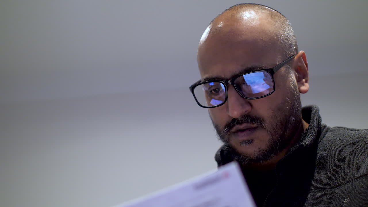 hombre calvo británico del sur de asia con gafas revisando las facturas del hogar pareciendo visiblemente ansioso