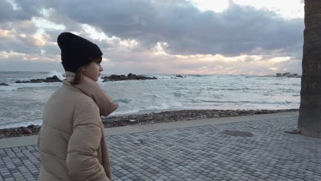 A-young-girl-walks-alone-along-the-boardwalk-with-angry-sea-waves-crashing-ashore-in-the-background-in-slow-motion