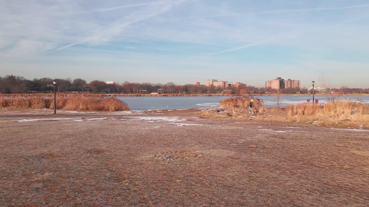 Aerial drone footage captures a man running through a park, passing by from a side angle as the camera smoothly transitions toward a frozen lake