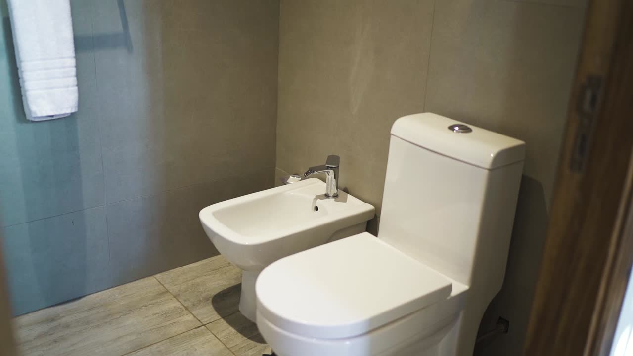 A modern hotel bathroom featuring a toilet and sink with clean, minimalist design and lighting, establishing tracking reveal right