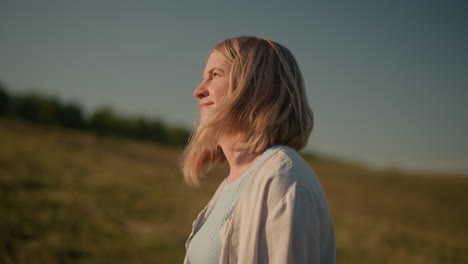 woman in blue top and light jacket walking outdoors with sunlight reflecting off her face, peaceful and relaxed, blurred background featuring scenic hilly farmland