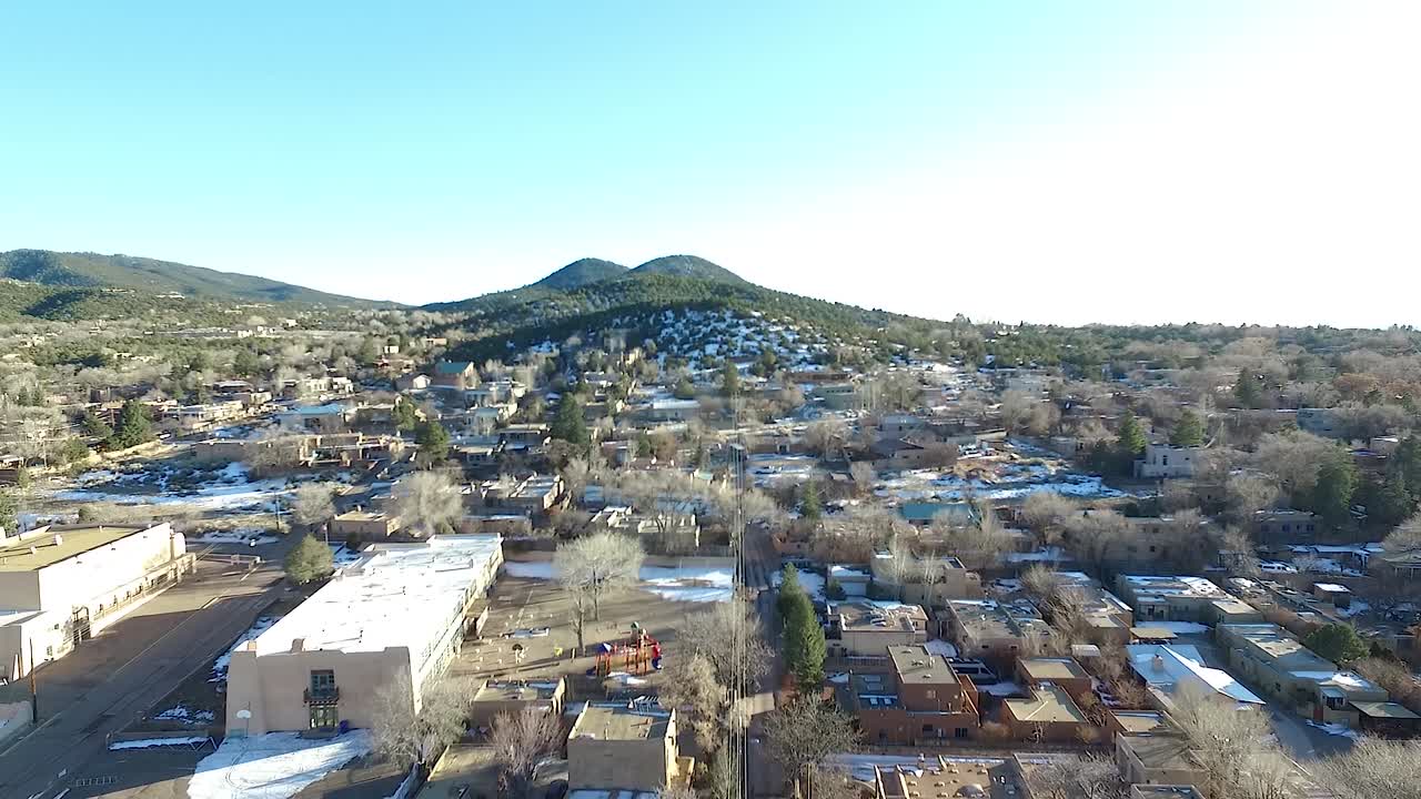 Aerial drone footage flying over a residential neighborhood in Santa Fe, New Mexico during winter. Snow patches are visible on rooftops and the ground, contrasting with distant green rolling hills