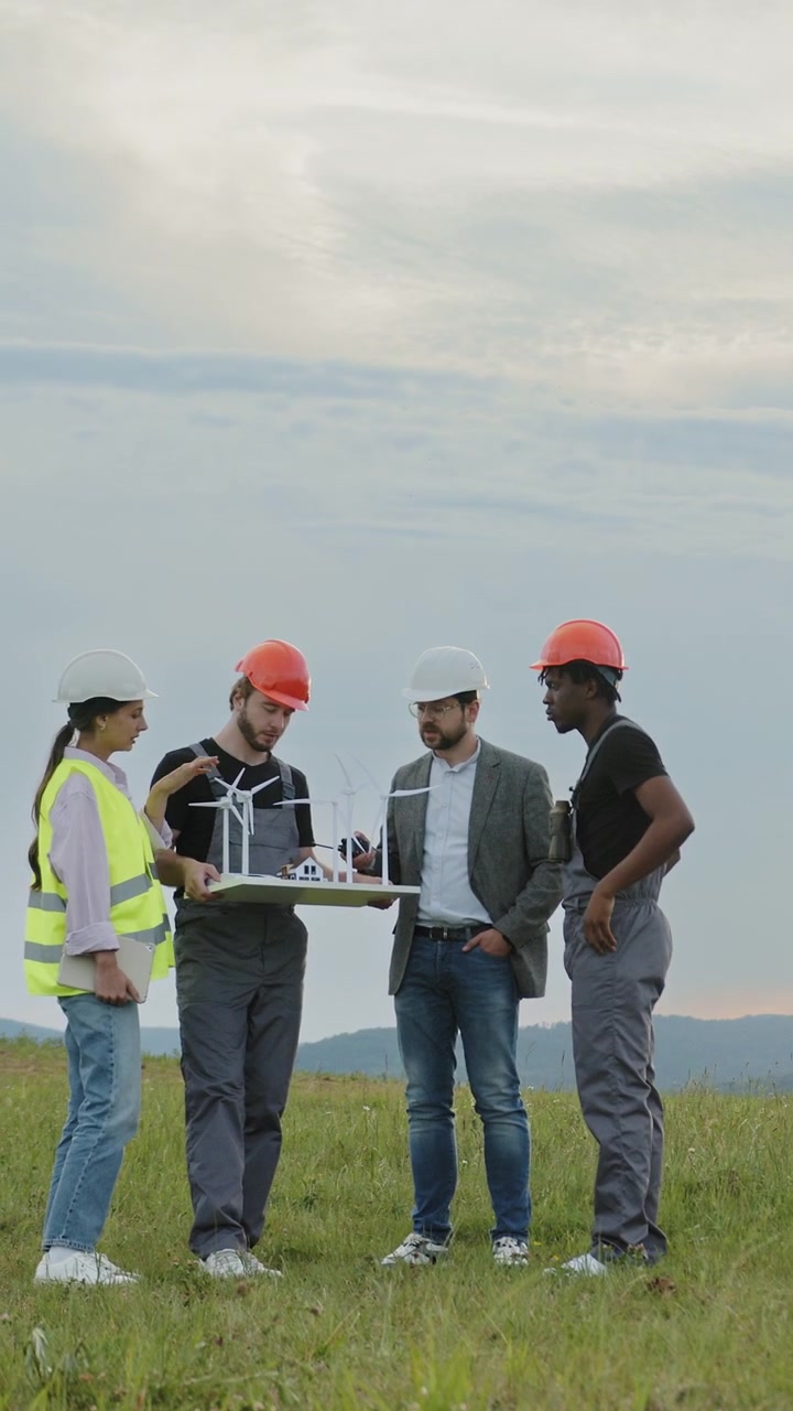 Engineers in hard hats and safety vests discuss a wind turbine model on a grassy field under a