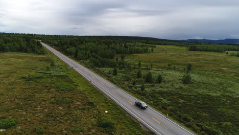 wide aerial shot of a veteran car driving on a long, straight country road surrounded by open green fields and patches of forest. northern mountain terrain in late summer.