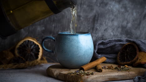 Pouring-fresh-herbal-tea-out-of-pot-into-blue-vintage-cup-decorated-with-ingredients-like-cinnamon,-dried-oranges-and-dried-tea-leaves