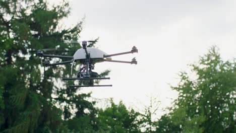 desde un ángulo bajo, una cámara de mano captura un avión no tripulado blanco y negro con seis hélices y una cámara, volando sobre un parque con árboles y arbustos