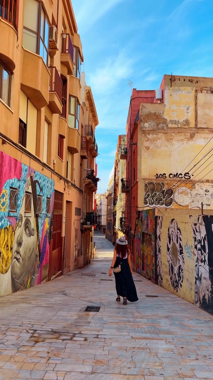 Alicante, Spagna, 8 agosto 2025: Donna che esplora un vicolo coperto di graffiti, Alicante, Spagna. Vista posteriore tra murales colorati su facciate consumate sotto una calda luce pomeridiana