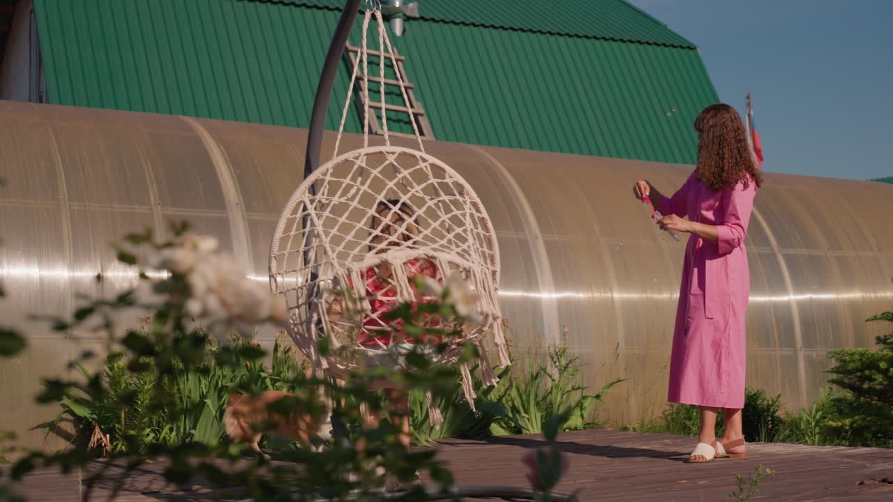 Girl Seated On Wicker Swing With Caregiver Nearby, Child Relaxes On Outdoor Swing While Caregiver Stands Close By, Young Girl Enjoys Sitting On Wicker Swing Surrounded By Garden Scenery And Pet Dog