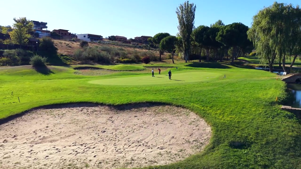 Tre mænd spiller golf på en solskinsdag og putter på en smuk green nær en sandbunker og en sø