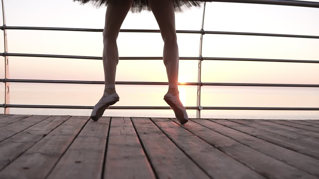 Aim footage of a ballet dancer's feet while she practices pointe exercises on the wooden embankment near sea. Close up of woman's legs in pointe shoes. Sunrise. Slow motion