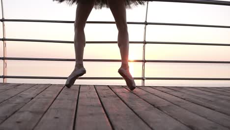 Aim-footage-of-a-ballet-dancer's-feet-while-she-practices-pointe-exercises-on-the-wooden-embankment-near-sea.-Close-up-of-woman's-legs-in-pointe-shoes.-Sunrise.-Slow-motion