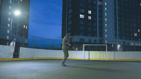 football player kicks soccer ball with precision toward goal during night training session on illuminated urban field, surrounded by tall residential buildings and vibrant blue evening sky