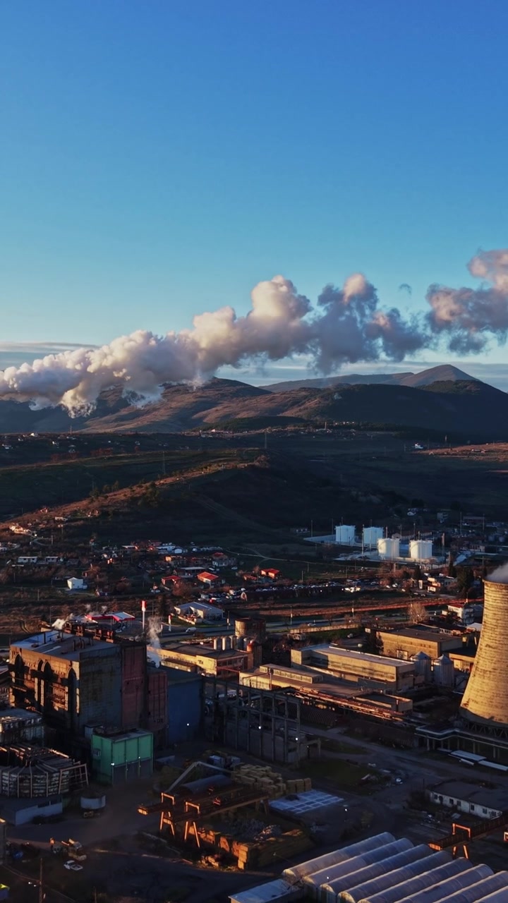 A fumaça sobe de edifícios industriais perto de montanhas ao pôr do sol