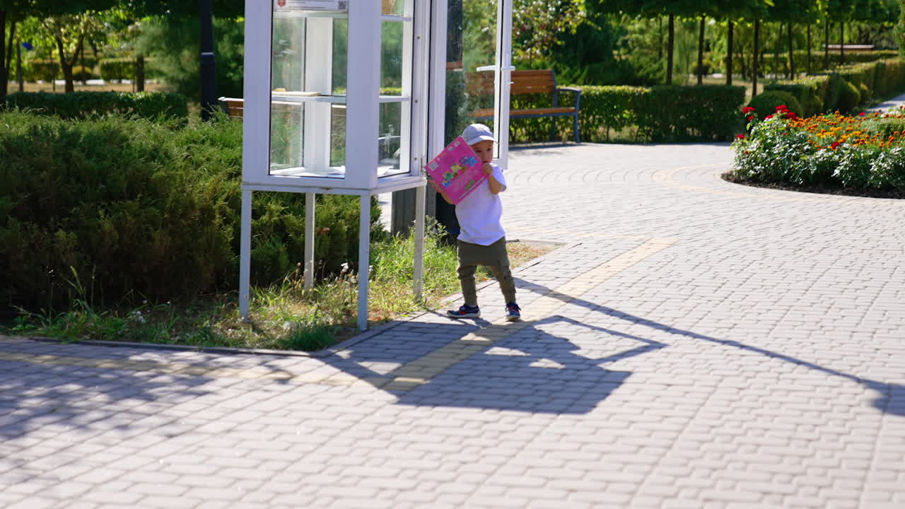 Cute little boy in a cap stands near the booth in the park in summer. Kid takes a pink magazine from the booth.