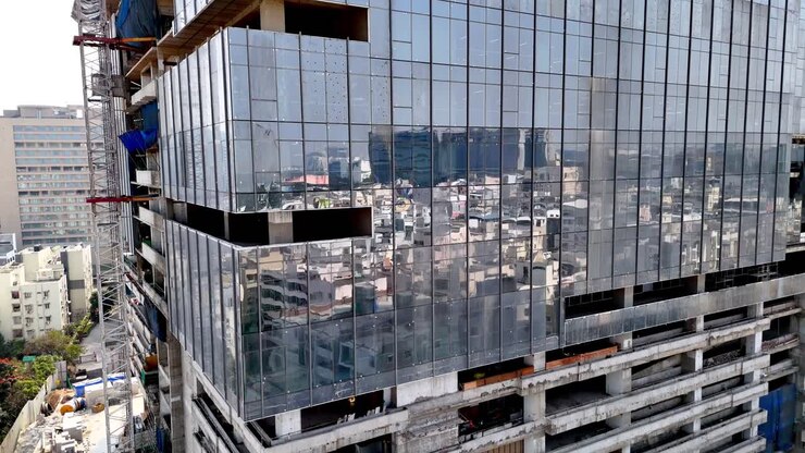 Vista aérea cercana de un rascacielos en construcción con fachada de cristal que se eleva bajo plantas de hormigón activas, andamios y trabajadores dando forma al perfil urbano de un distrito de negocios urbano de rápido crecimiento.