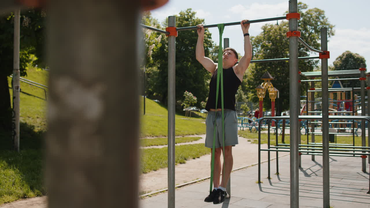 Athletic man in sportswear doing pullups exercises on horizontal bar with help of elastic expander
