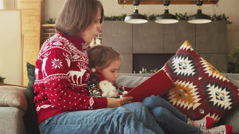 encantadora madre sentada en el sofá con una hija adorable y leyendo su cuento de hadas el día de navidad en casa