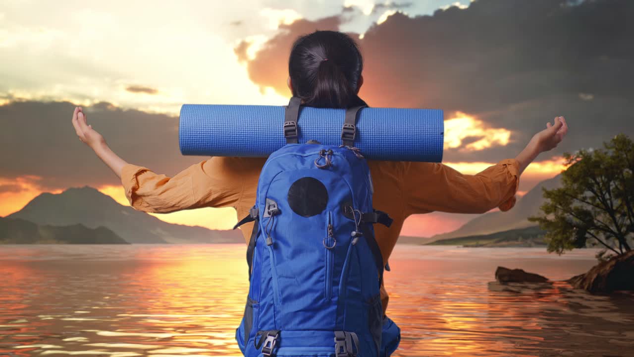 vista trasera de una excursionista femenina con mochila de montañismo extendiendo los brazos y mirando la vista a su alrededor mientras está de pie en un lago durante la hora del atardecer