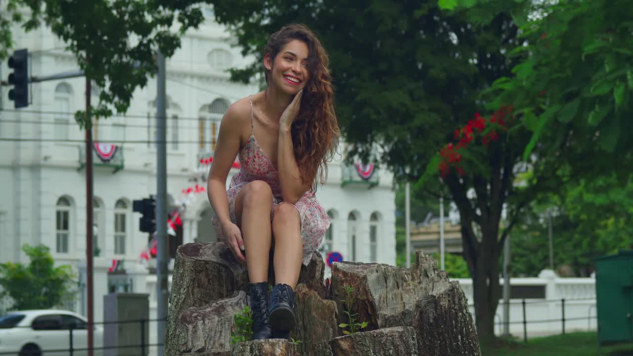Bella latina con capelli ricci siede su un ceppo d'albero e sorride in minidress con un castello sullo sfondo in un parco.