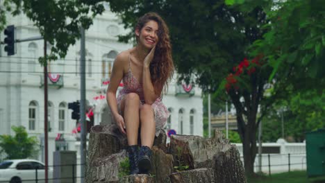 hermosa latina de cabello rizado se sienta en un tronco de árbol y sonríe en un vestido mini con un castillo en el fondo en un parque