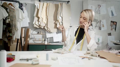 Mujer-Sastre-Sentada-En-La-Mesa-Eligiendo-Piezas-De-Ropa-Y-Hablando-Por-Teléfono-En-El-Taller