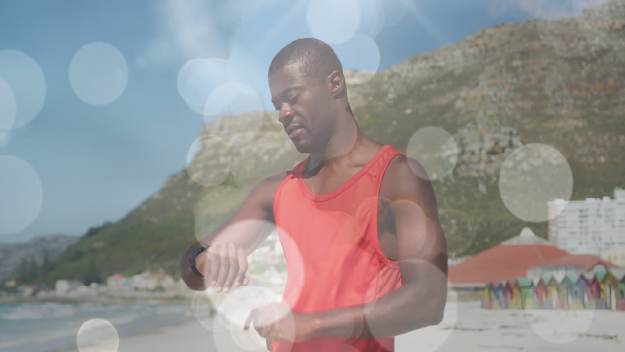 animación de luces brillantes sobre un hombre afroamericano entrenando en la playa usando un reloj inteligente