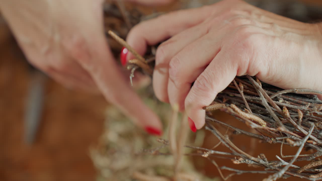 Female florist hands with red nails gripping bundle of twigs while crafting rustic wreath on wooden table, highlighting detail of handmade process, artistic creativity decoration work