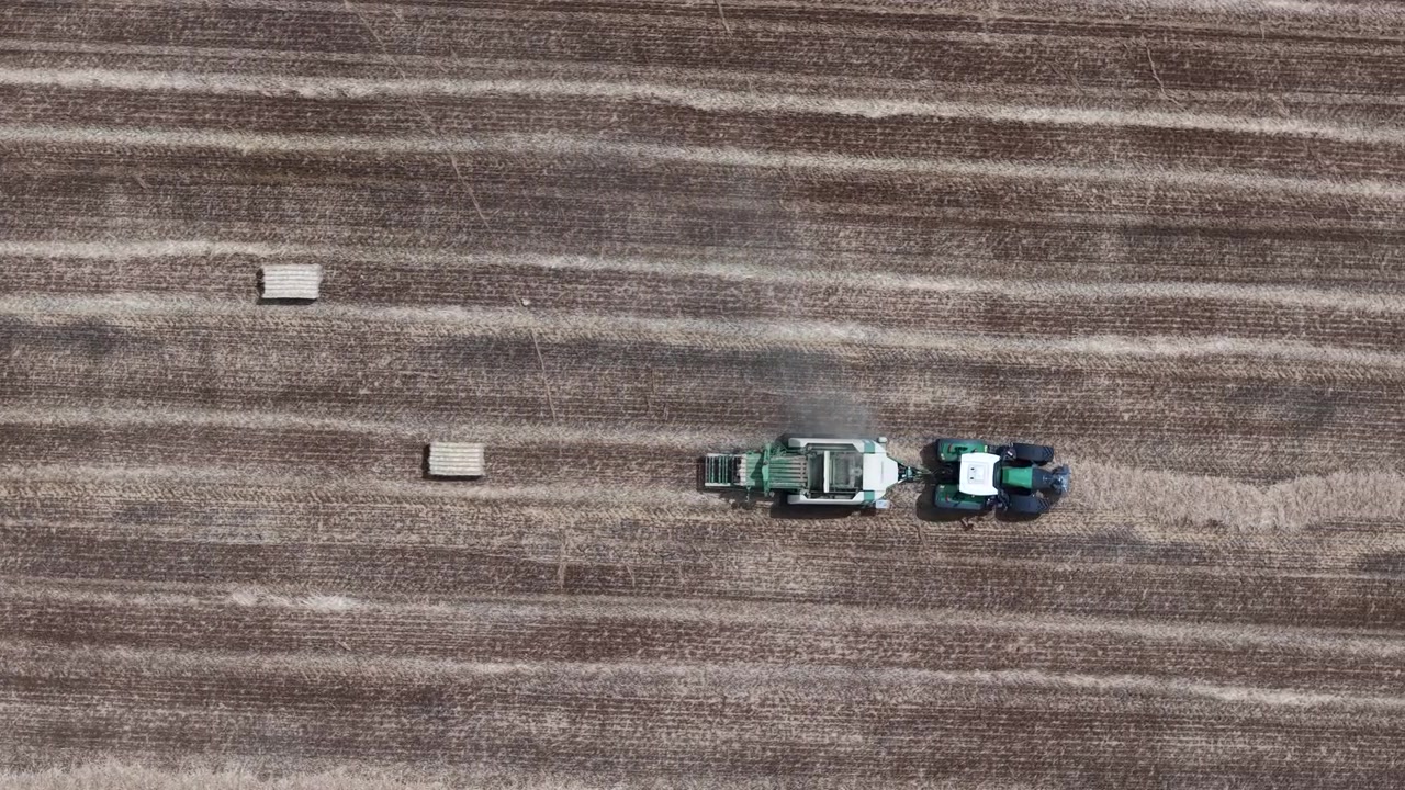 Una vista aerea mostra un trattore che traina una pressa per balle, creando balle di paglia in un vasto campo agricolo durante la stagione del raccolto.