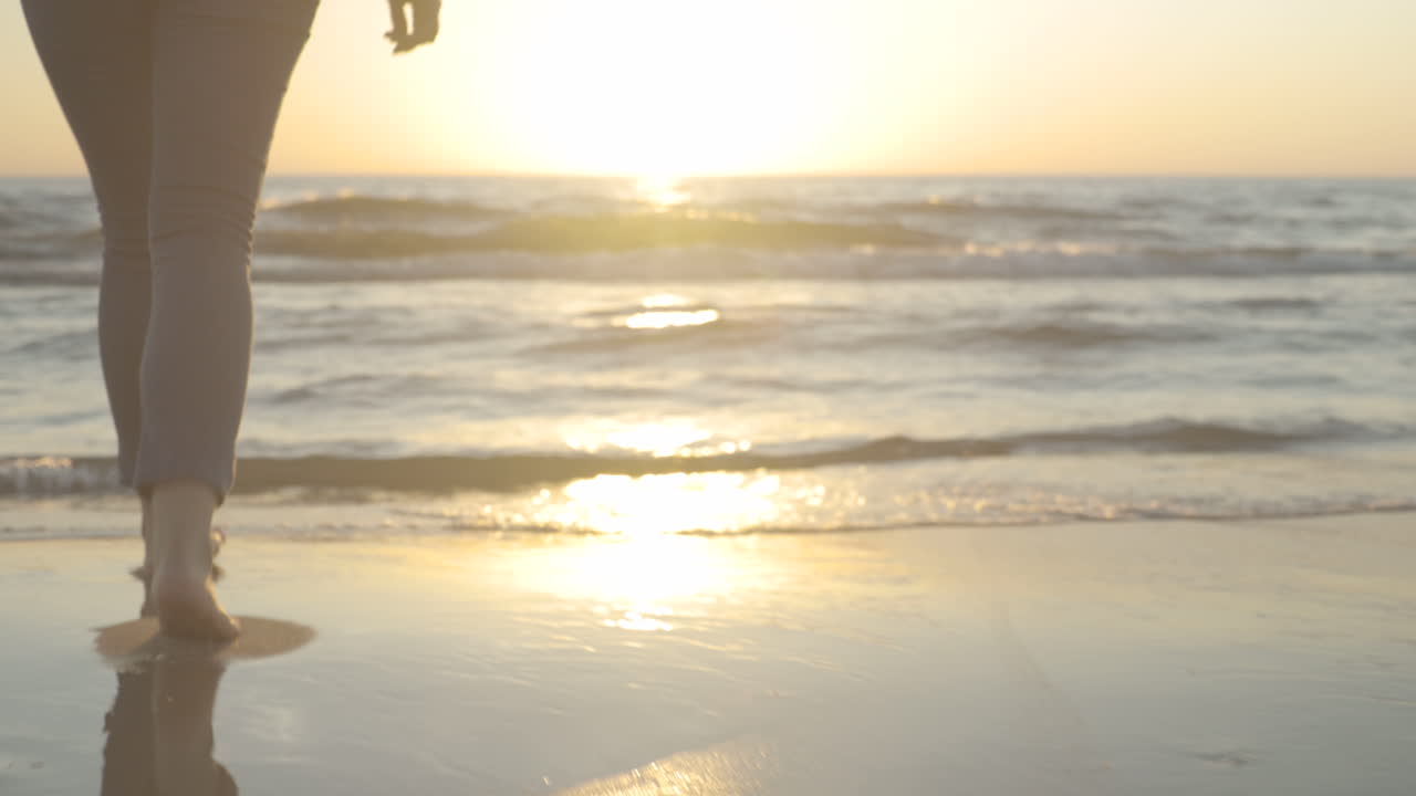 Handheld shot of walking feet on a beach.  Walking toward the water.  Backlit by the setting sun.  Shot in 4K