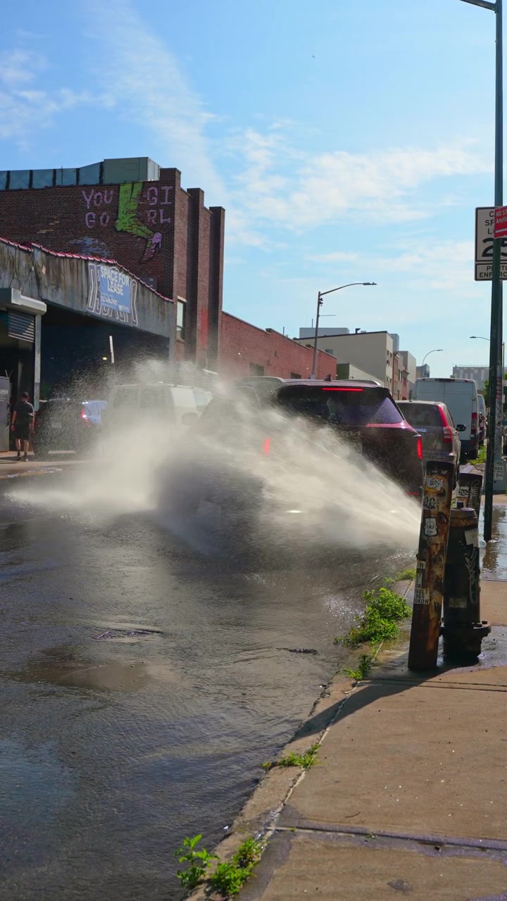 New York, USA, 5 giugno 2026: Idrante che spruzza acqua su una strada cittadina industriale. Asfalto bagnato che scorre oltre pali e furgoni alla luce del sole.