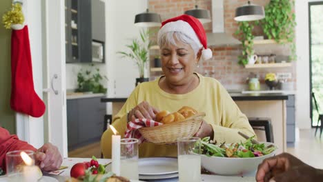 group of happy diverse senior friends in santa hats passing food at christmas dinner table at home