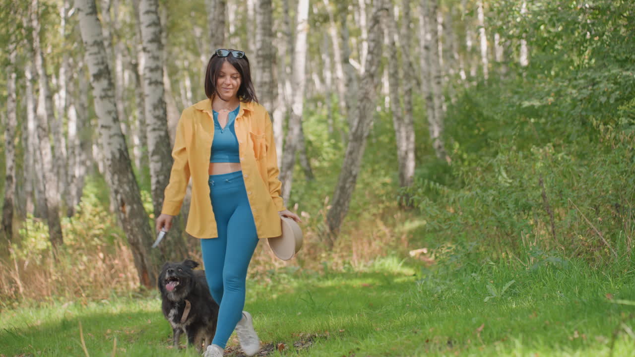 Black woman walking birch forest with dog in yellow jacket and teal outfit holding hat, upbeat stroll along sunlit trail, playful pup at side, lively connection and relaxed camping vibe
