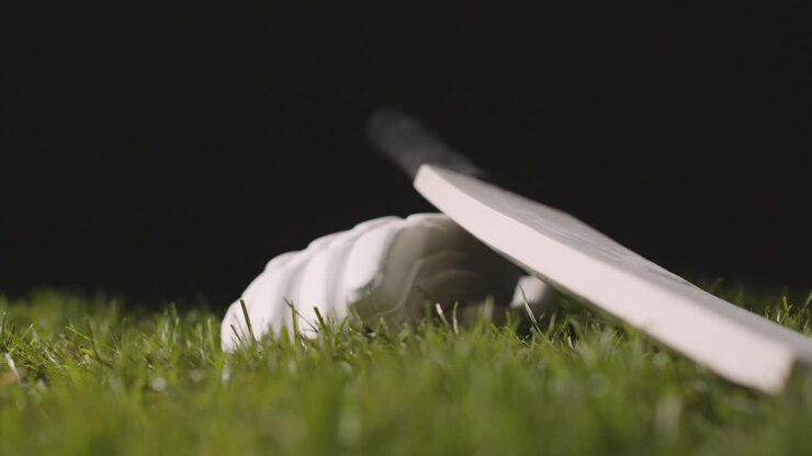 Natura morta di cricket in studio con primo piano di una palla che rotola accanto a mazza e guanto adagiati sull'erba 1.