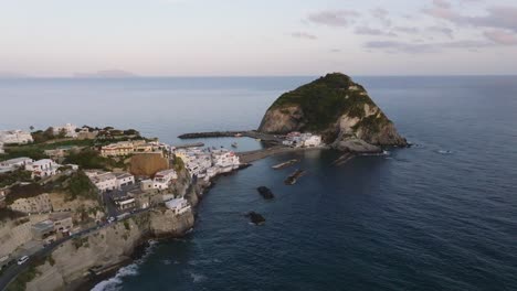 Houses-by-cliffy-shore,-islet-and-open-ocean-at-Ischia-at-dusk,-aerial