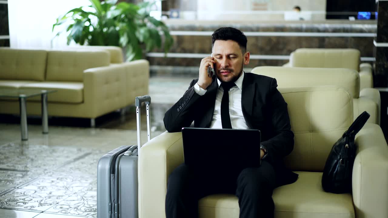 hombre de negocios atractivo y confiado hablando por teléfono móvil y usando una computadora portátil mientras se sienta en un sillón en un hotel de lujo con una maleta de equipaje cerca de él. concepto de viaje, negocios y personas