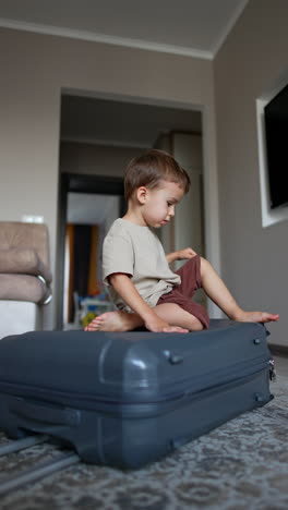 lovely little boy sits on the big grey suitcase lying on the floor. kid puts his foot on the wheel of the suitcase. vertical video.