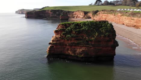 aerial drone flyover of the clear and tranquil waters with a grass-covered, impressive rock along the cliffside of ladram bay