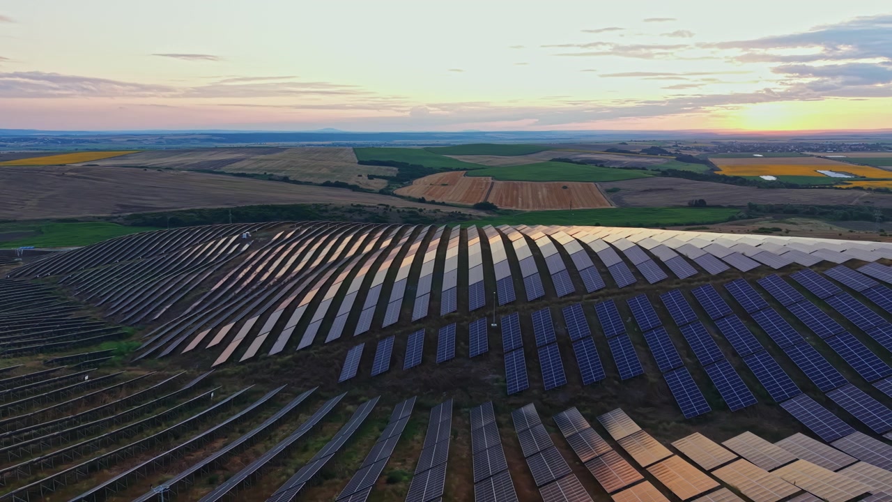 Solpaneler dækker et stort felt under solnedgang nær et landområde med afgrøder og landbrugsjord i det fjerne