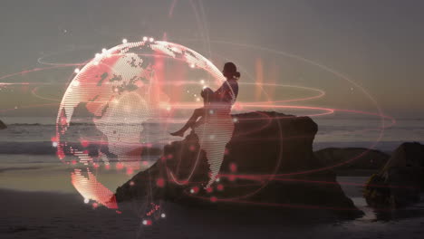 woman using handheld device on rocky beach at dusk, with glowing digital globe technology overlay