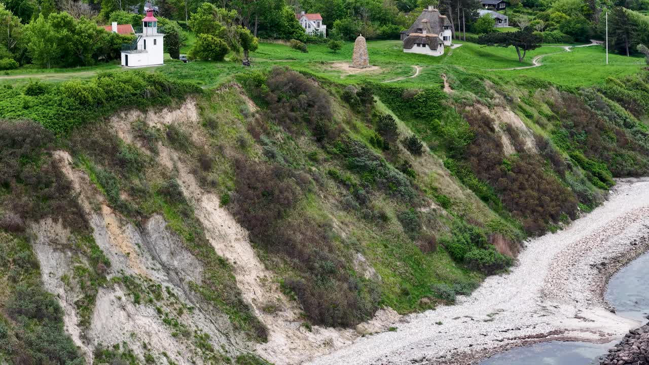 Drone footage showcasing a steep grassy cliff on the coast of Denmark. The aerial view captures eroded slopes, green vegetation and a narrow sandy beach where gentle waves lap the shore