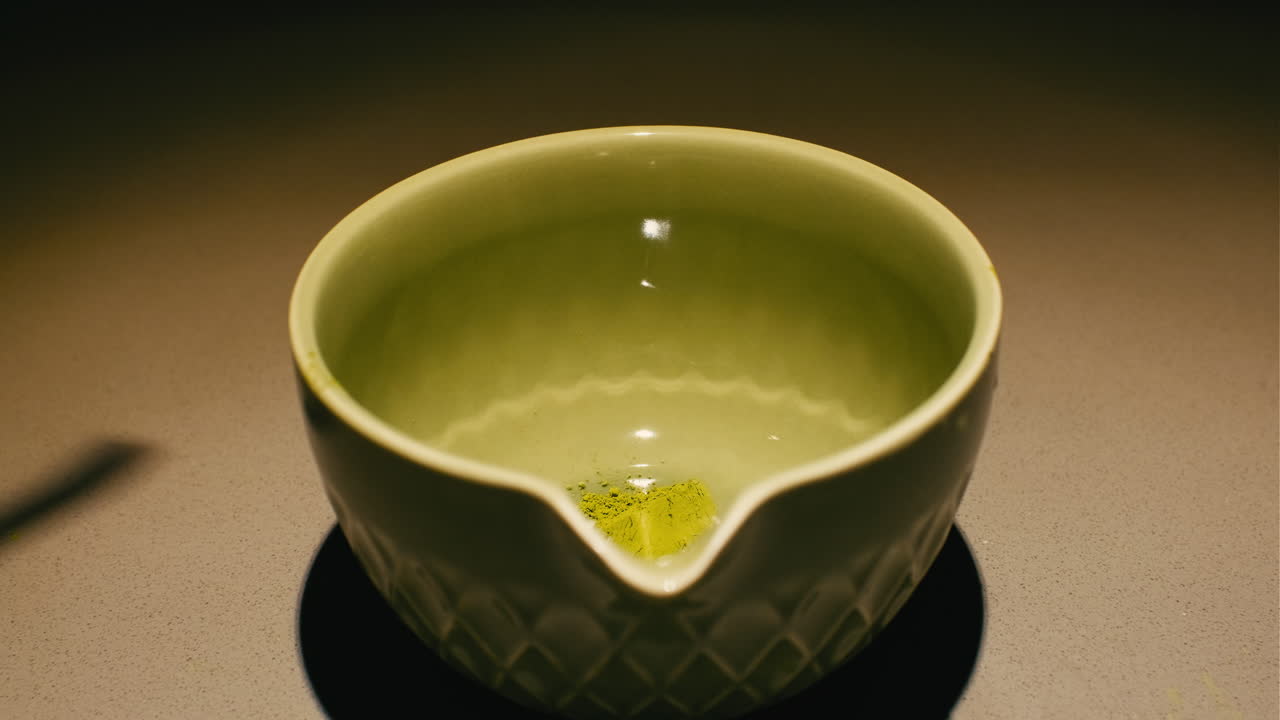 Closeup of green tea powder being added to a ceramic bowl and mixed with hot water, perfect for wellness and culinary content