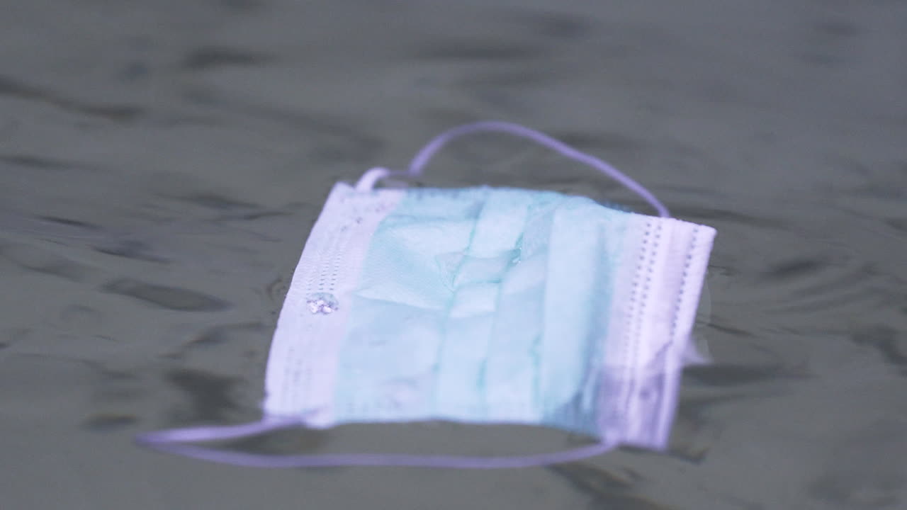 회색 구름과 비오는 날 연못의 불안한 물 위에 떠 있는 단순한 녹색 보호용 코비드-19 얼굴 마스크, 누군가에 의해 버려지고, 흠뻑 젖고, 정적 클로즈업 4k 샷