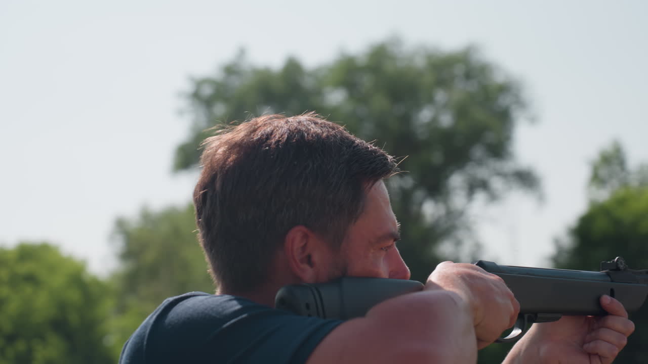 close up side view of man aiming rifle with focused gaze outdoors on sunny day, beard visible, tension in posture, background blurred trees and sky, preparation for pest control or protection