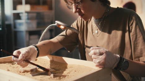 female archaeologist using brush to clean bones in sand-filled box