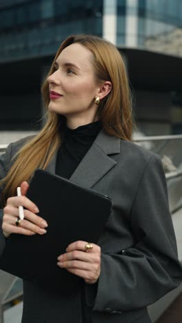 businesswoman architect in a gray suit stands on a modern office terrace, holding a tablet and stylus, working, writing, or brainstorming, smiling in admiration of the building she designed