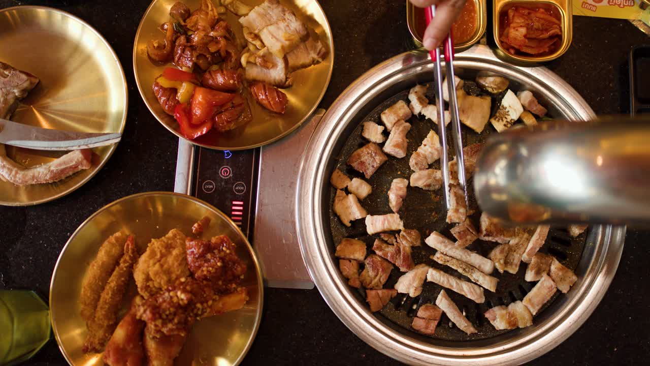 Hand uses tongs to turn pork belly pieces on a smoking tabletop grill, overhead view