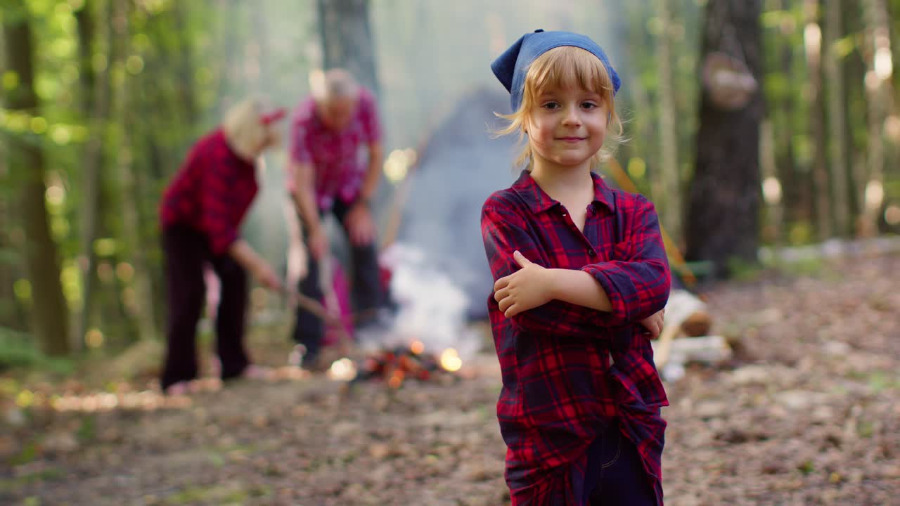 Child girl kid traveler tourist on camping looking at camera showing thumbs up near bonfire in wood