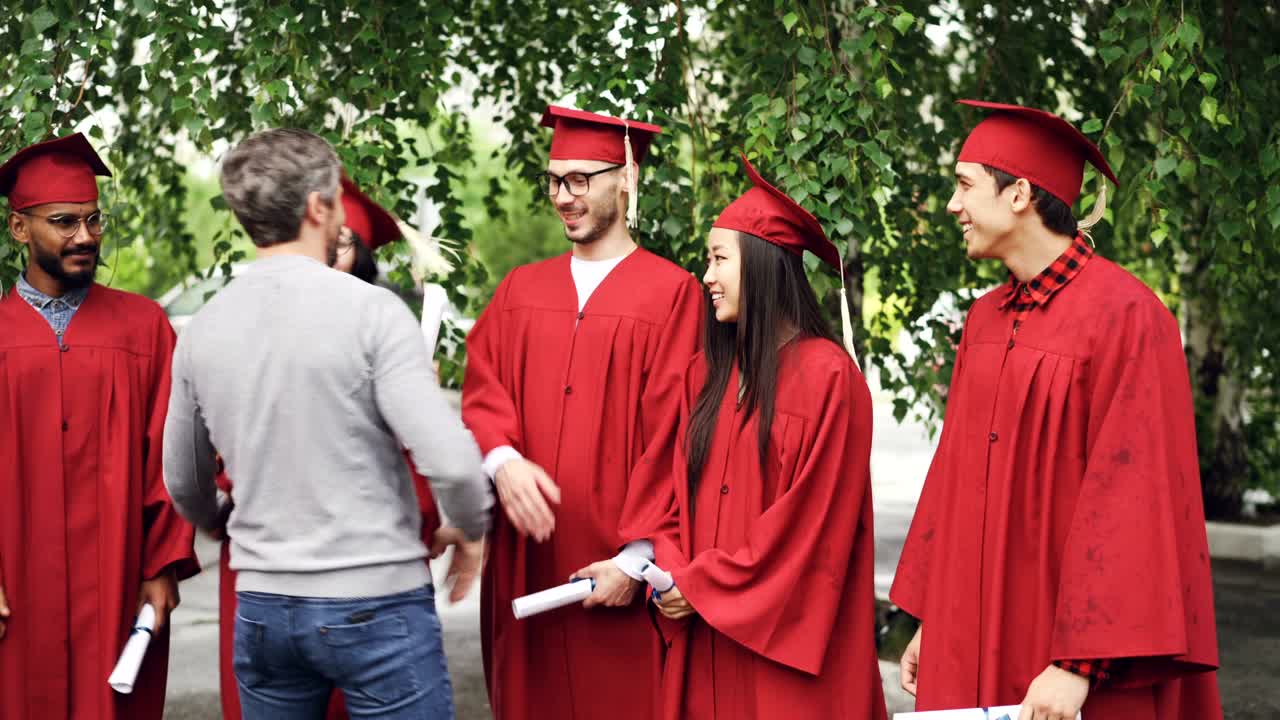 Bearded man proud teacher is congratulating graduates shaking hands and hugging them outdoors in college campus while students are talking and holding diplomas.